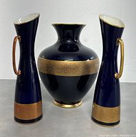 View of the full set showing two tall narrow pitchers flanking a wide rounded vase. All pieces are navy blue with gold decorative bands and gold handles on the pitchers.