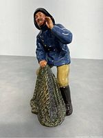 Front view of the Royal Doulton Sea Harvest figurine showing the fisherman holding a green fishing net, wearing a blue coat, cream pants, brown boots and black hat.