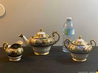 Lidded teapot, sugar bowl with lid, and creamer displayed beside water bottle for scale on black tablecloth