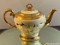 Front-angle view of golden hand painted teapot showing the handle, body with floral design, and part of the spout.