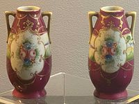 Pair of antique Nippon porcelain vases shown front with visible floral hand painted designs on burgundy background, gold accenting and dual handles, about 5 inches high.