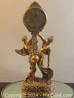 Front view of the vintage gold-tone mantle clock showing two dancing ladies holding the clock face with Roman numerals.