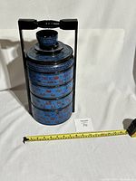 Full view of the four-tier lacquer tiffin with handle, showing overall shape and size with visible tape measure for scale.