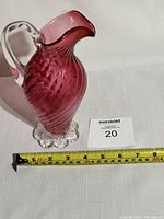 Photo of cranberry glass pitcher with tape measure showing width of about 5 inches and height of about 8 inches, placed on white background with MaxSold lot number card.
