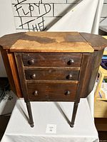 Full front view of the antique sewing cabinet showing three drawers, drop-leaf sides in upright position, and wear on the top surface.