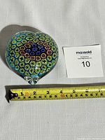 Top view of heart-shaped glass paperweight showing the details of the millefiori design and measuring tape indicating size.