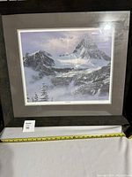 Full view of framed print showing mountain scene under glass with dark frame and measuring tape below for size reference.