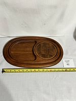 Wooden teak cutting board, oval shape with two recessed compartments, measuring tape showing approx 15 inches length.