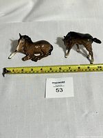 Two porcelain horse figurines placed on white fabric with measuring tape, one lying and one standing.