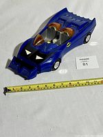 Blue vintage toy Batmobile car photographed from an angle showing front-left side, with a tape measure for scale execution around 12 inches long.
