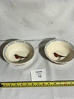 Two ceramic bowls shown from top angle, each with robin bird design and 'ROBIN' text around rim. Measuring tape indicates size about 19 cm wide.