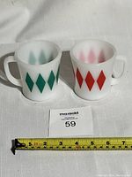 Pair of Fire King coffee cups with green and red Harlequin diamond patterns sitting on white fabric with measuring tape showing about 3.5 inches tall.