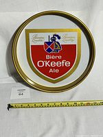Top down photo of the circular metal sign featuring the O'Keefe Ale logo, with measuring tape indicating size.