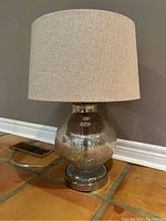Single round silver textured table lamp with beige fabric drum shade placed on a tiled floor against a neutral wall.