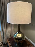 First round silver gloss finish table lamp with white shade placed on wooden side table next to curtains