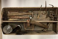 Full view of wooden toolbox with all tools inside showing variety of antique and vintage hand tools including hammers, hooks, saw blade, metal rake tool, glass jar, and others.