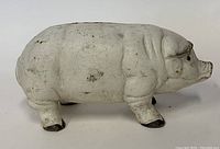 Side view of the white cast iron piggy bank shaped like a pig with black hooves and wear visible on surface.