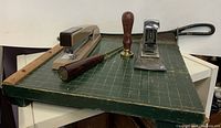 Photo showing a collection of antique and vintage office items on a green cutting mat including two staplers, a wax seal with wooden handle, a metal paper punch or cutter with black handle, and a wooden ruler.