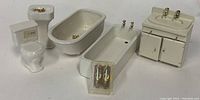 Photo depicting five pieces of white miniature bathroom furniture: toilet, pedestal sink, clawfoot bathtub, bathtub with taps, and bathroom vanity with double sinks and cabinet doors. Small chandelier light fixture in plastic case included in front.