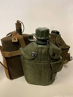 Photo showing three military canteens side by side: WWII vintage with brown wool cover and cork stopper, 1950s style green metal canteen with olive drab nylon cover, and smaller canteen with tan canvas cover