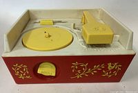 Front angle view of vintage Fisher Price red and white toy record player with yellow turntable and tonearm. Shows decorative floral and bird motifs and volume control knob.