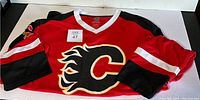 Red Calgary Flames hockey jersey with flaming 'C' logo, black and white sleeve accents, and V-neck collar with tags.