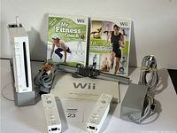 Full lot overview showing Nintendo Wii console upright on stand, 2 Wiimotes, power adapter, sensor bar, Wii Operations Manual, and 2 fitness games.