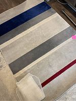 Photo of the rectangular rug showing blue, grey, white and red horizontal stripes along its length with non-slip liner partially visible on one edge.
