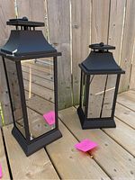 Image showing two black metal outdoor lanterns with clear glass panels, set on a wooden deck next to a wooden fence.
