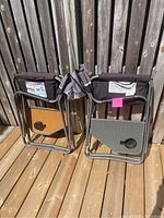 Two folded Coleman camping chairs leaning on a wooden fence with visible foldable side tables on each chair.