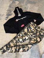 Full view of the black Champion hoodie with logo and camouflage Adidas pants laid flat on floor.