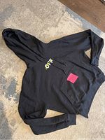 Black Off-White hoodie showing front with small yellow graphic and front pocket spread on carpet with red tag on chest.
