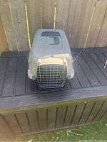 Front view of light gray and beige hard case pet carrier with metal mesh front door, placed on dark surface outdoors.