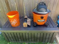 Ridgid shop vac with orange bucket and small box on a brown plastic deck box lid outdoors