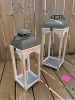 Two lanterns standing upright on a wooden deck showing the wooden frame and metal tops.