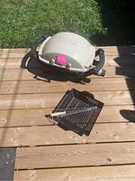 Small light gray Weber BBQ grill placed on wooden deck with perforated vegetable grilling tray and two metal skewers.