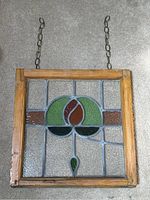 Full view of the antique stained glass window showing the wooden frame, hanging chain, and central floral motif in green, red, brown, and black glass.