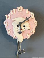 Front view of the pink circular clock face featuring a sculpted white cat, raised numerals, and dark gray hands.