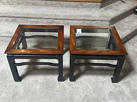 Pair of square vintage end tables shown from front, with black legs and warm brown wooden edges around beveled glass tops on carpeted floor.