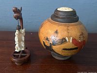 Miniature ivory or bone figure hanging on a dark wooden stand with dragon carving next to a round painted jar with Asian figures