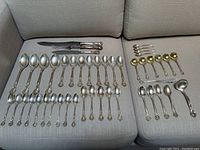 Overall view of utensils arranged on sofa, showing carving knives, spoons, small spoons, mustard spoons, butter knife, ladle