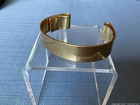 Gold tone cuff bracelet displayed on a clear acrylic stand on a blue cloth background, showing side and top angles.