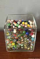 Front view of the clear square glass container filled with various marbles of different colors and sizes.