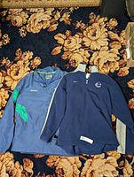 Two garments laid flat on carpet: blue Reebok jacket with green accents and navy-blue Nike Canucks sweater with grey sleeves