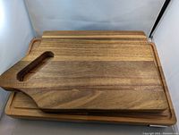 Two wooden cutting boards stacked. Smaller board has handle with oval cutout, placed on top of larger board with juice groove.