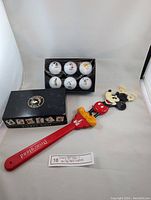 Photo of 6 white golf balls with Disney character prints inside a black Disney Golf branded box and a red Disneyland Mickey Mouse back scratcher.