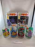Photo showing two Funko Pop figures of Grimace and Hamburglar in boxes and four vintage McDonald's collectible glasses featuring characters Ronald McDonald, Birdie, Hamburglar, and Grimace.