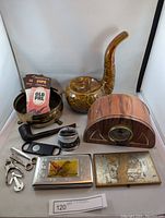 View of full lot including ceramic pipe ashtray, vintage pipe fillers, pipe tools, vintage cigarette cases, wooden mantel clock, and assorted pipe accessories.