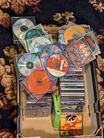 Tray filled with CDs, some loose discs on top, others stacked in jewel cases showing various artist labels and covers.