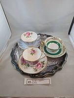 Photo shows three vintage bone china teacups with floral designs (pink roses and green band), each with matching saucers, arranged on a copper plated scalloped tray with reflective surface and signs of light wear.